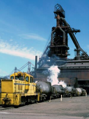 "A yellow locomotive and its line of special-purpose wagons waits in front of a blast furnace as molten metal is poured into a wagon in the background.  Steam rises from the hot, grimy containers."